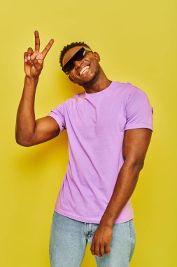 Mutlu Afro-Amerikan adam son moda yaz kıyafetleri ve güneş gözlükleriyle Sarı üzerinde zafer işareti gösteriyor.
