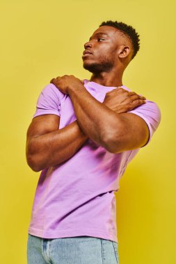 Kendine güveni olan, mor tişörtlü Afro-Amerikan adam sarı arka planda poz veriyor ve başka yöne bakıyor.