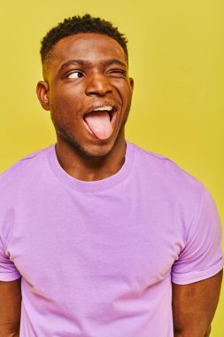 Komik Afro-Amerikalı mor tişörtlü adam dilini dışarı çıkarıyor ve sarı arka plana bakıyor.