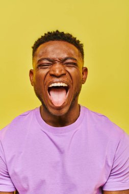 Genç Afro-Amerikalı, gözleri kapalı, yüzünü buruşturan ve sarı arka fonda dilini dışarı çıkaran adam.