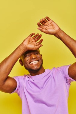 Aşırı neşeli ve şık Afro-Amerikan erkeği mor tişörtlü sarı arka planda gülüyor.