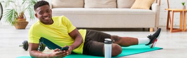 smiling black man with smartphone looking at camera on fitness mat near sports bottle, banner clipart