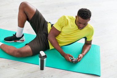 Genç atletik Afrikalı Amerikalı adam spor şişesinin yanındaki spor minderinde akıllı telefondan mesajlaşıyor.