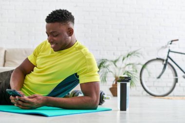 Akıllı telefonlu gülümseyen Afrikalı Amerikalı adam spor şişesinin yanındaki spor minderinde internette geziniyor.