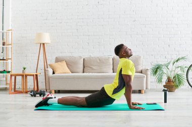 Spor kıyafetli Afro-Amerikan bir adamın evde spor minderinde yılan yogası pozu çalışmasının yan görüntüsü.
