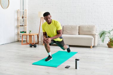 sporty african american guy working out on fitness mat near sports bottle and smartphone at home clipart