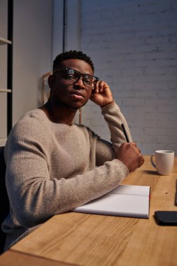 Gözlüklü Afrikalı Amerikalı adam ofisteki not defterinin yanındaki kameraya bakıyor, serbest çalışan.