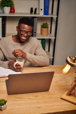 Kahve fincanı taşıyan neşeli Afro-Amerikalı adam ev telefonunda gülüyor, serbest çalışan.