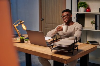 joyful african american man with coffee cup smiling during video call in home office, freelancer clipart
