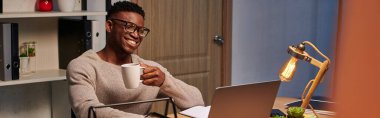 positive african american freelancer with coffee smiling during video chat in home office, banner clipart