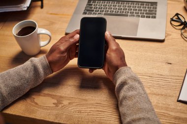 Afro-Amerikan serbest çalışanın elinde bilgisayarın yanında boş ekranla akıllı telefon tutuşu görüntüsü.