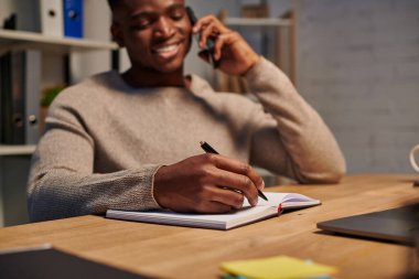 Afro-amerikan serbest çalışan mutlu akıllı telefondan konuşan ve not defterine yazan, evden çalışan