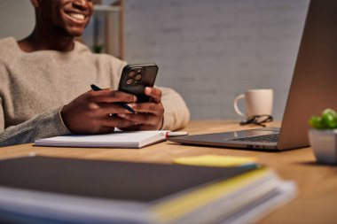 Gülümseyen Afrikalı Amerikalı adam akıllı telefondan mesaj atıyor. Evdeki dizüstü bilgisayarın yanında, kırpık manzaralı.