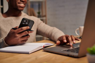 Akıllı telefonlu mutlu Afro-Amerikan adam evde çalışırken dizüstü bilgisayar kullanıyor.