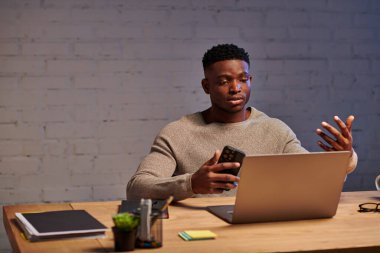 Genç Afrikalı Amerikalı serbest çalışan. Akıllı telefonlu. Evdeki bilgisayardan yapılan video görüşmesi sırasında el kol hareketi yapıyor.