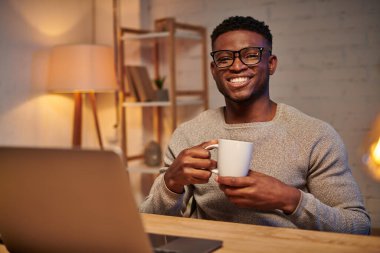 Keyifli Afro-Amerikan serbest çalışanı kahve fincanı ile gecenin bir yarısı merkez ofiste dizüstü bilgisayarın yanında oturuyor.