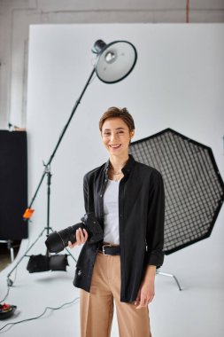 Kamerasıyla poz veren ve neşeyle gülümseyen kısa saçlı neşeli genç bayan fotoğrafçı.