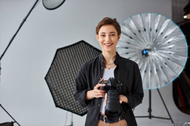Kamerasıyla poz veren ve mutlu bir şekilde gülümseyen kısa saçlı neşeli genç bayan fotoğrafçı.