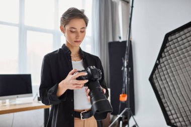 Günlük kıyafetli genç ve çekici bir kadın fotoğrafçı stüdyosundaki fotoğraf makinesine bakıyor.