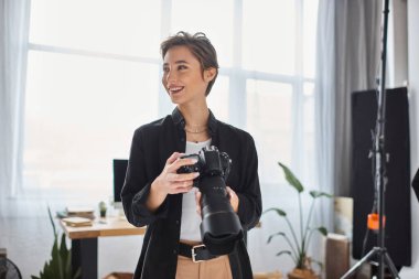 Neşeli kadın fotoğrafçı, günlük giysiler içinde, elinde kamera, gülümsüyor ve başka tarafa bakıyor.