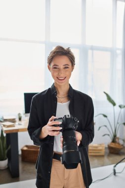 Kamerayla poz veren ve neşeyle gülümseyen kısa saçlı kadın fotoğrafçı.