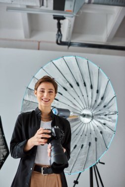 Kamerayla poz veren ve neşeyle gülümseyen kısa saçlı kadın fotoğrafçı.