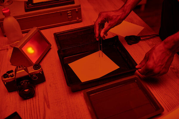 cropped shot of man delicately immerses photo paper in a darkroom bath with chemicals, red light