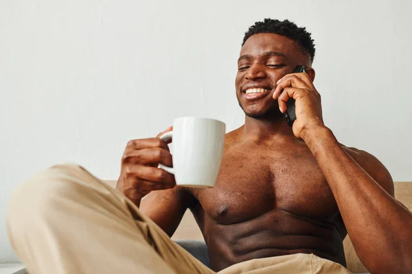 Stock image cheerful muscular african american man with morning coffee talking on smartphone in bedroom