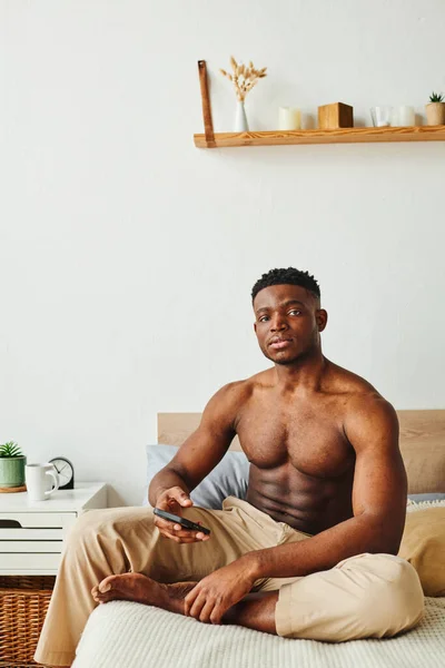 muscular african american man in pajama pants sitting on bed with smartphone and looking at camera