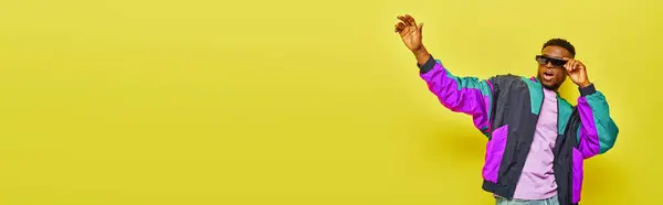 stock image thrilled african american man in sunglasses and bright jacket waving hand on yellow backdrop, banner