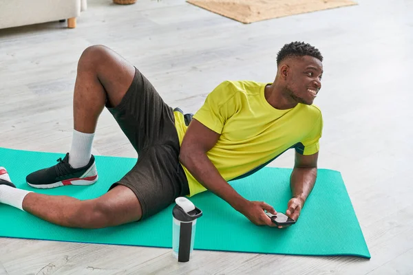 快乐运动的非洲男人 拿着手机 在运动瓶子旁边的健身垫上遥望着 — 图库照片