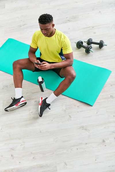 身穿运动服的非洲裔美国人 在运动瓶子和哑铃附近的健身垫上拿着智能手机 — 图库照片