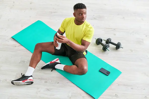 身穿运动服的年轻的非洲裔美国人坐在健身垫上 旁边的运动瓶靠近智能手机 — 图库照片