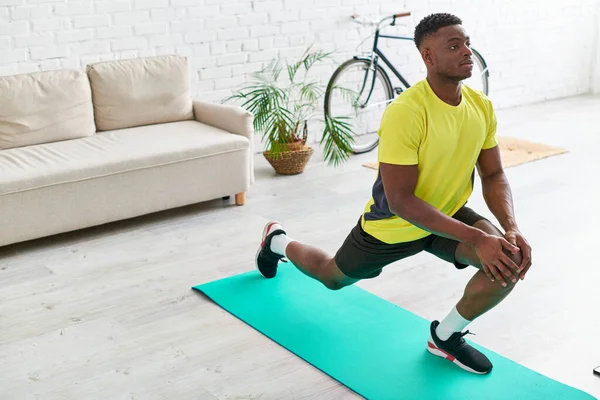身穿运动服的英俊的非洲裔美国男子在家中客厅的健身垫上伸懒腰 — 图库照片