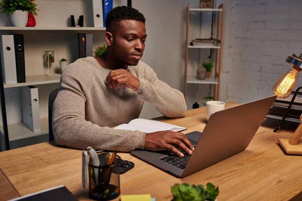 concentrated african american man typing on laptop while working in evening at home, freelancer