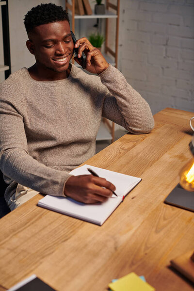 веселый африканский американец разговаривает на смартфоне рядом с ноутбуком и ноутбуком во время работы из дома