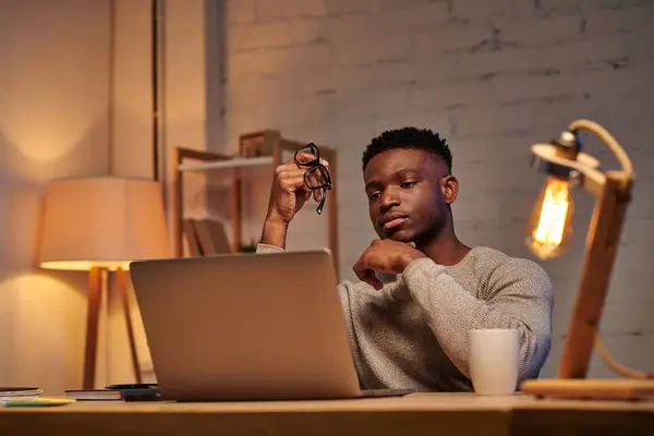 Stock image thoughtful african american freelancer holding eyeglasses looking at laptop in home office at night