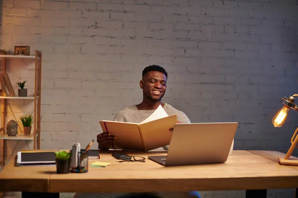 joyful african american freelancer with notebook looking at laptop while working at home at night