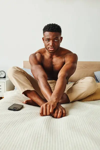 confident and strong african american man in pajama pants sitting on bed and looking at camera