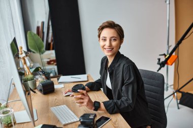 happy attractive female photographer holding camera while retouching photos and smiling at camera clipart