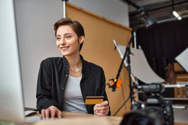 Neşeli, çekici kadın fotoğrafçı kredi kartını kullanarak internetten ödeme yapıyor ve mutlu bir şekilde gülümsüyor.