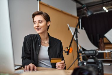 cheerful attractive female photographer paying online using her credit card and smiling happily clipart