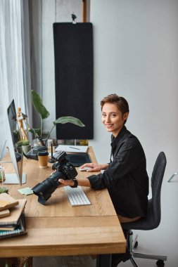 joyous charming female photographer smiling at camera while working on her photos in studio clipart