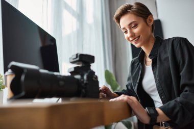 Tablet ve fotoğraf makinesiyle bilgisayarın önünde çalışan neşeli, güzel bir kadın.