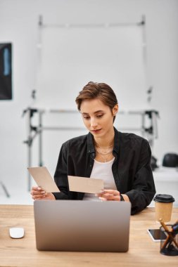 beautiful female photographer in casual attire sitting at table and looking at photos in her hands clipart