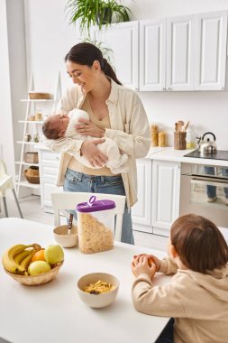 jolly attractive mother enjoying breakfast with her toddler and newborn sons, modern parenting clipart