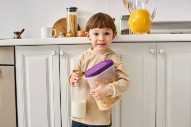 adorable toddler boy in cozy homewear holding milk and corn flakes during breakfast on kitchen clipart