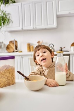 cute toddler boy in comfy homewear sitting at table during breakfast and looking at camera clipart