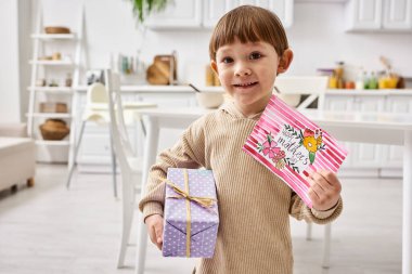Anneler Günü 'nde hediye ve tebrik kartıyla poz veren rahat ev elbiseli sevimli çocuk.