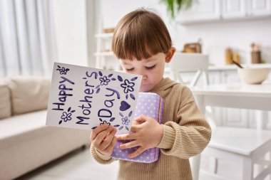 Anneler gününde hediye ve tebrik kartıyla poz veren rahat ev elbiseli sevimli küçük çocuk.